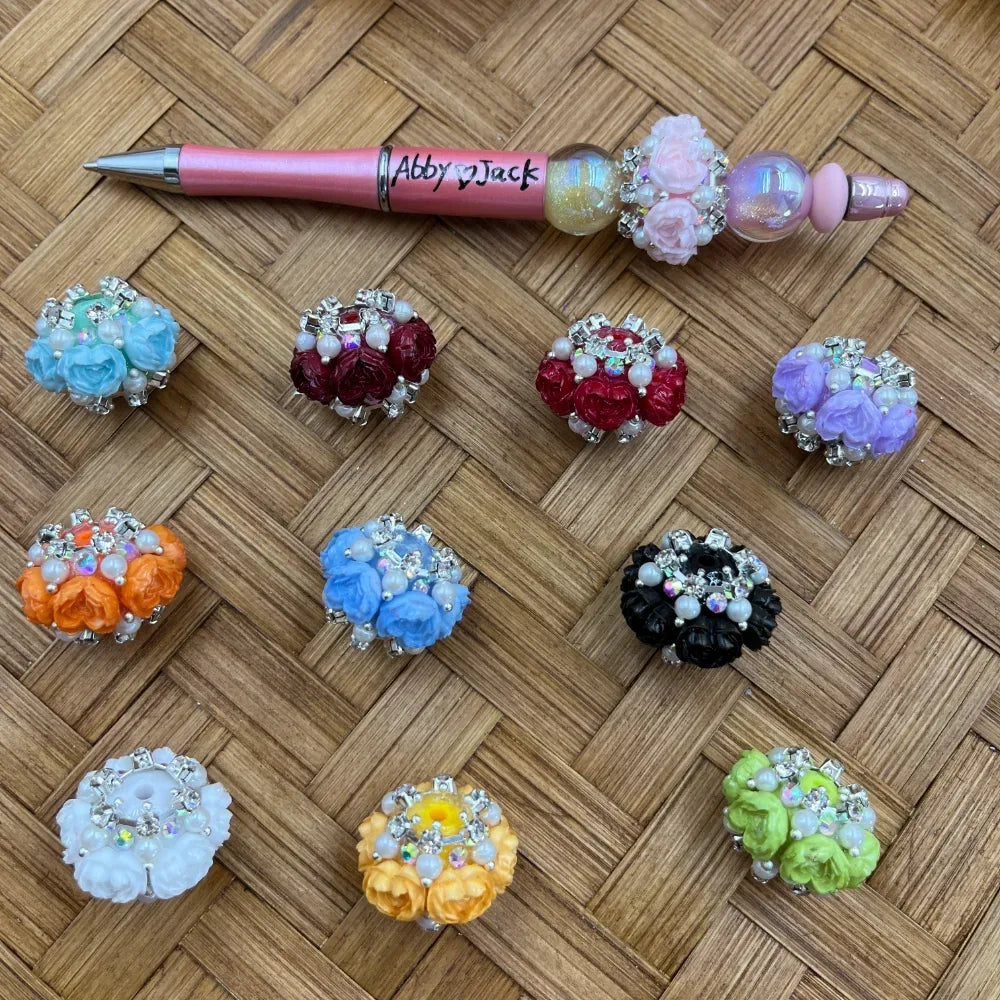 Assorted rhinestone rose cluster fancy beads in multiple colors including red, blue, orange, purple, black, white, yellow, and green with pearl and crystal accents, displayed alongside a decorated beadable pen on a wicker background.
