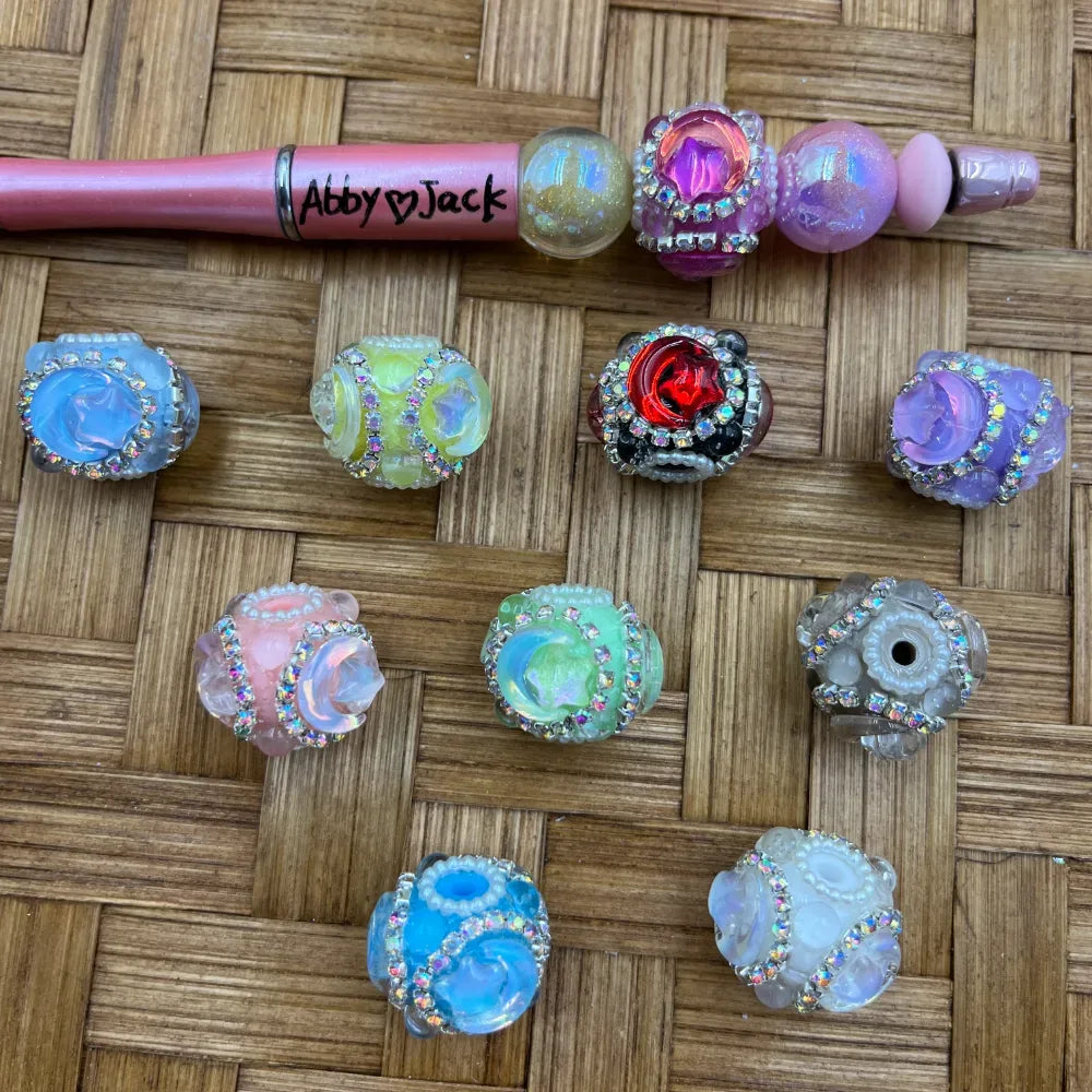 Assorted rhinestone-trimmed rose floral resin fancy beads in blue, pink, green, purple, red, teal and white opal colors displayed on wicker surface alongside a decorated beadable pen labeled Abby and Jack.