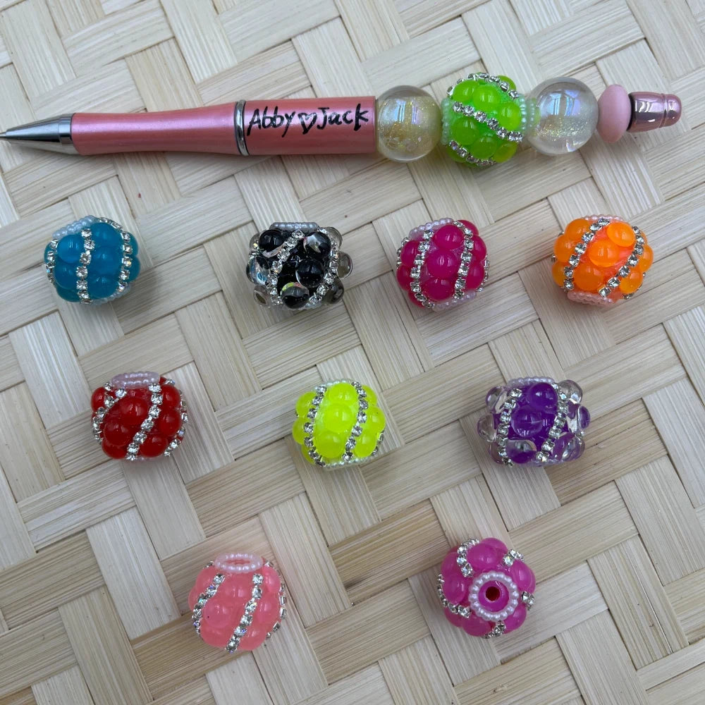 Assorted chunky bubbly rhinestone stripe fancy beads in teal, black, hot pink, orange, red, yellow-green, purple, coral, and magenta, displayed next to a beaded pen on a woven background.