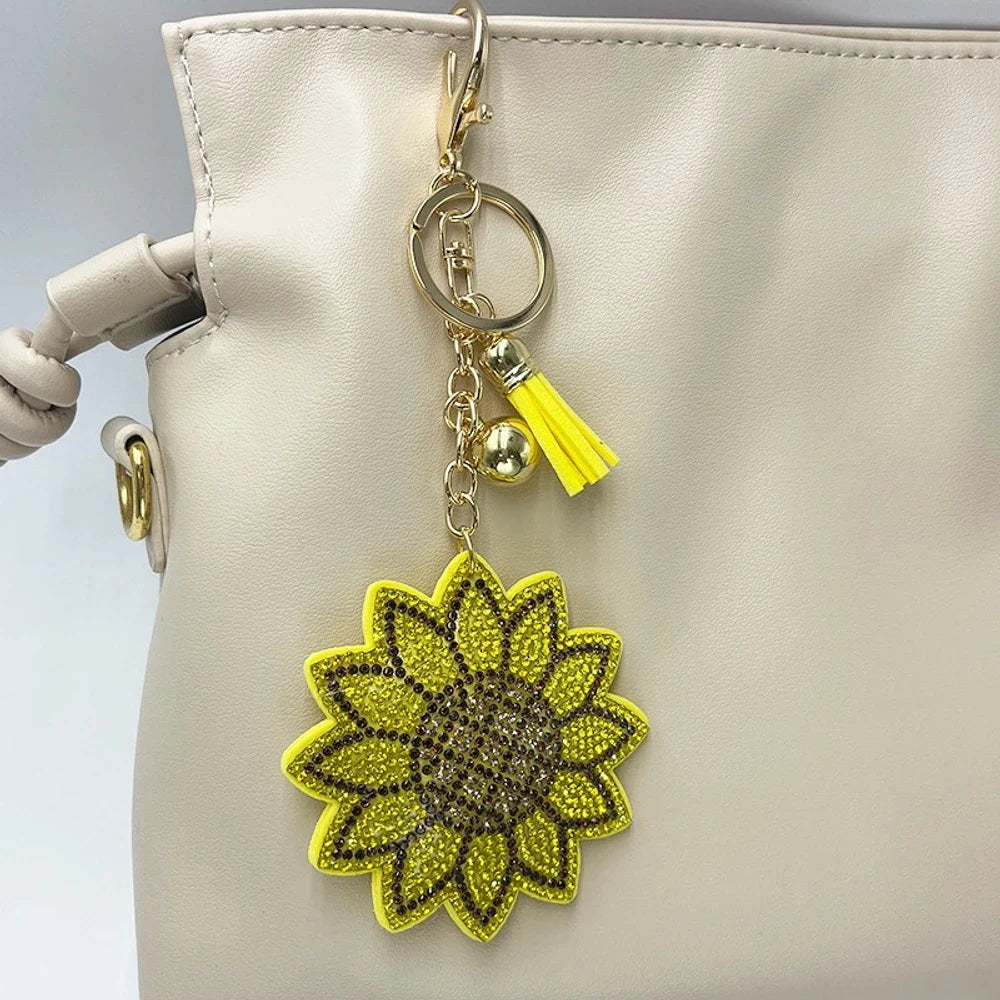 Yellow rhinestone sunflower keychain charm with gold lobster clasp, chain, gold beads, and yellow tassel hanging on a cream leather handbag.