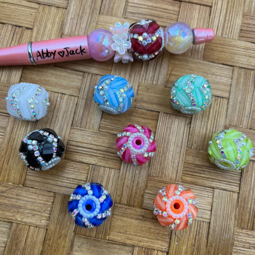 Assorted colorful rhinestone swirl chunky resin fancy beads in white, black, blue, pink, green, teal, and orange, displayed on a woven surface next to a decorated beadable pen labeled Abby Jack, featuring crystal AB rhinestone trim and pearl accents.
