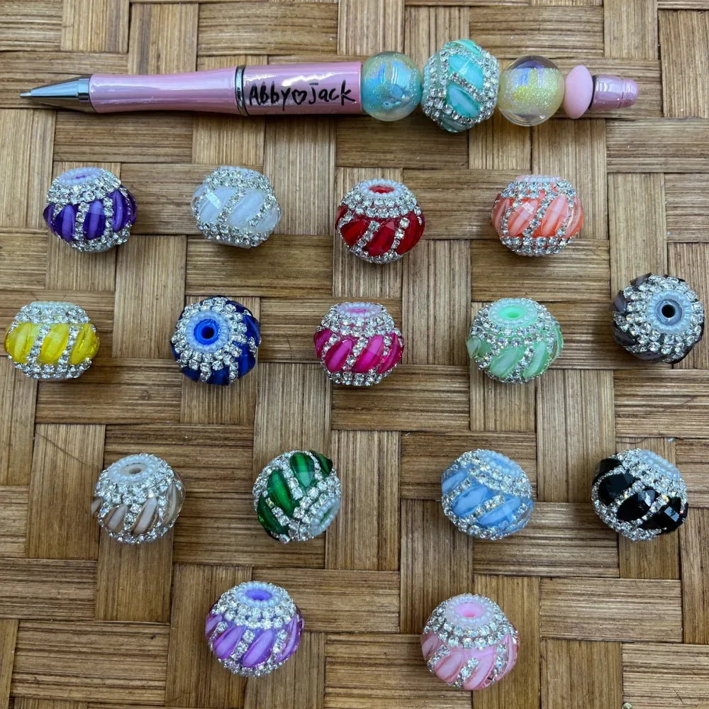 Assorted rhinestone-trimmed swirl striped round fancy beads in multiple colors including purple, white, red, orange, yellow, navy, hot pink, mint green, black, green, blue, lavender, and pink displayed on a woven surface next to a beadable pen.