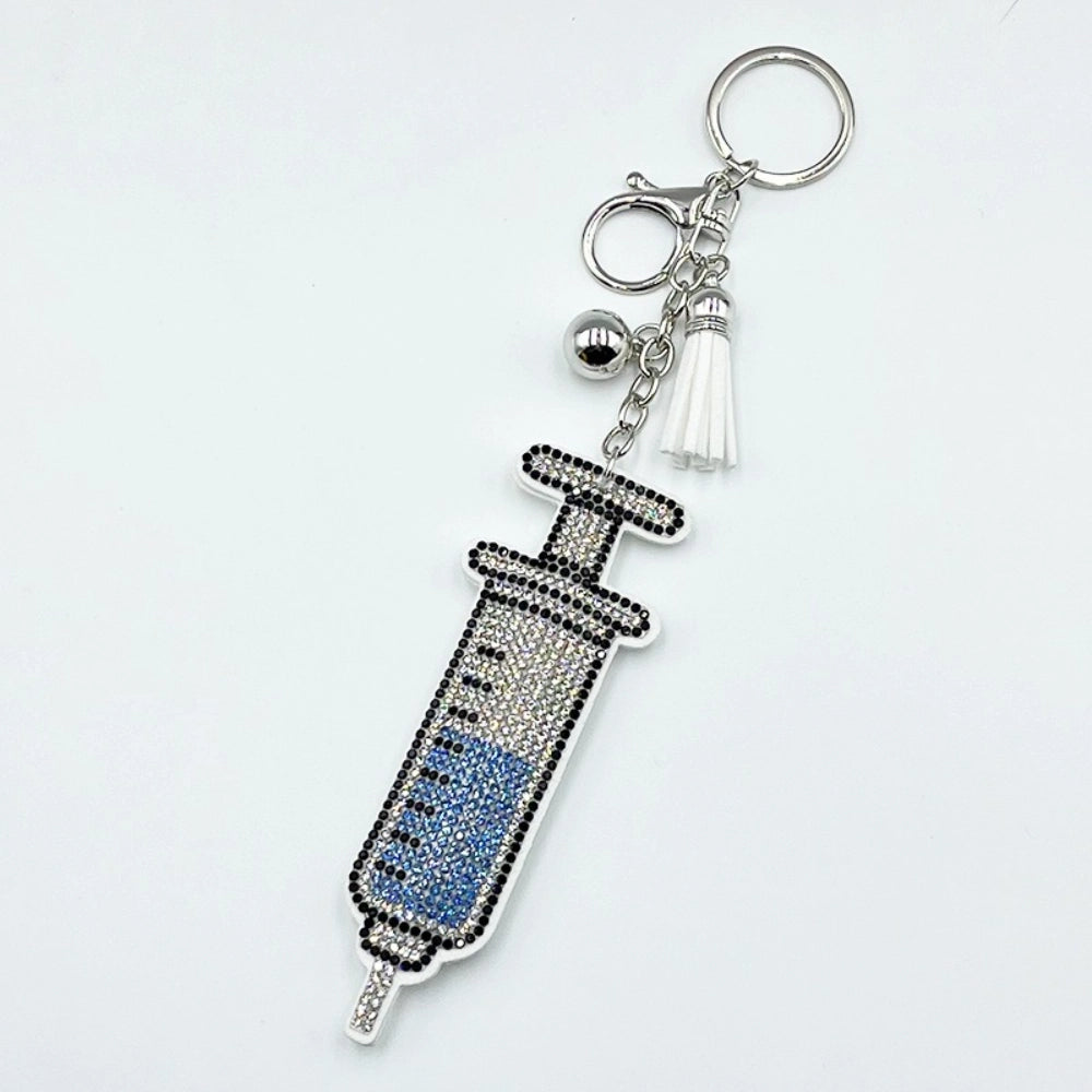 Crystal rhinestone syringe-shaped keychain charm with black and blue gemstones, white tassel, silver bell, and lobster clasp on white background.