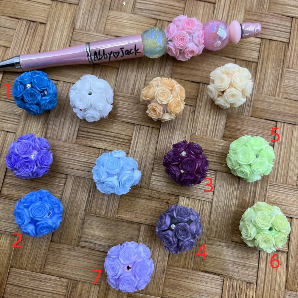 Colorful rose flower ball fancy beads in blue, white, peach, cream, purple, lavender, light blue, dark purple, green, and lilac displayed on a woven surface next to a decorative beadable pen labeled Abby and Jack.