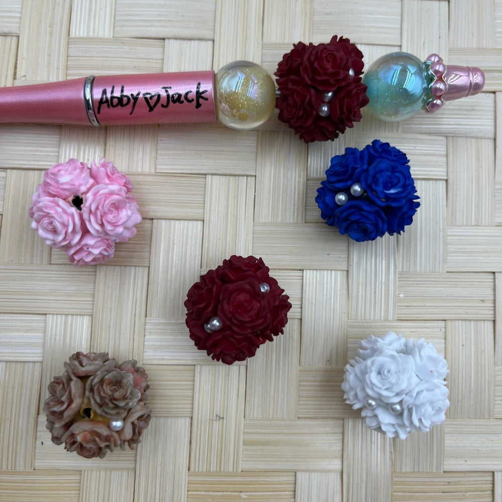 Assorted rose flower cluster fancy beads in burgundy, royal blue, pink, white, and taupe with pearl accents, displayed next to a custom beadable pen on a wicker surface.