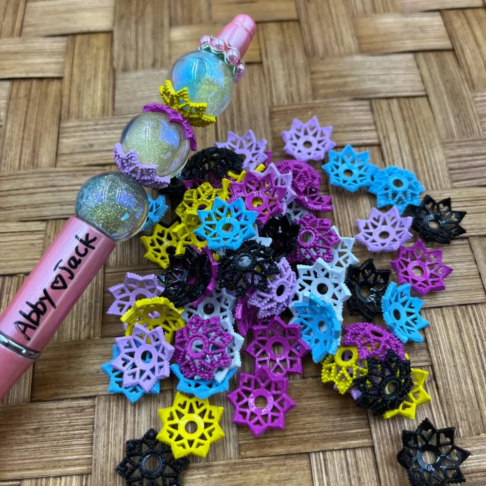 Colorful snowflake lace flower bead caps in purple, blue, yellow, black, and pink spread on a woven surface next to a decorated beadable pen labeled Abby and Jack with glitter globe beads.