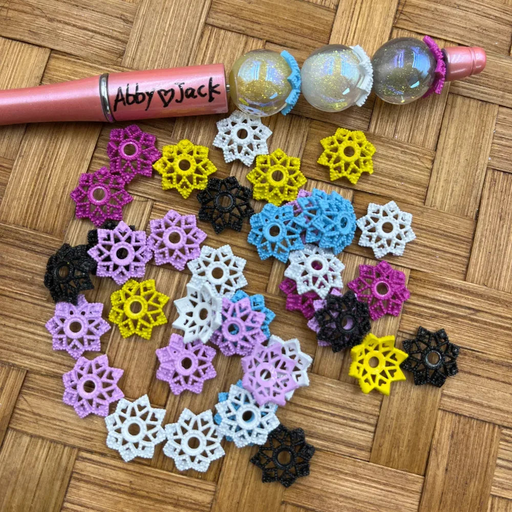 Assorted colorful snowflake lace plastic bead caps in pink, yellow, white, black, blue, and lavender spread on a woven surface next to a beadable pen and glitter globe beads.