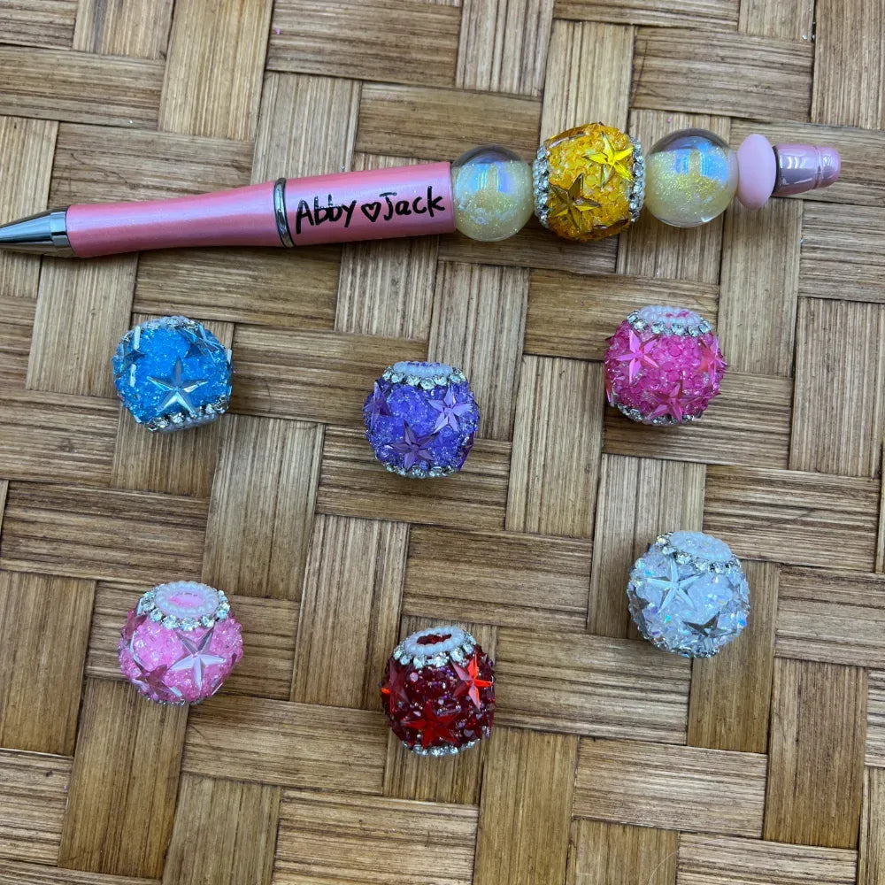 Colorful star crystal rhinestone fancy beads in blue, purple, pink, red, yellow, hot pink, and white, displayed next to a pink beadable pen labeled Abby and Jack on a woven bamboo surface.