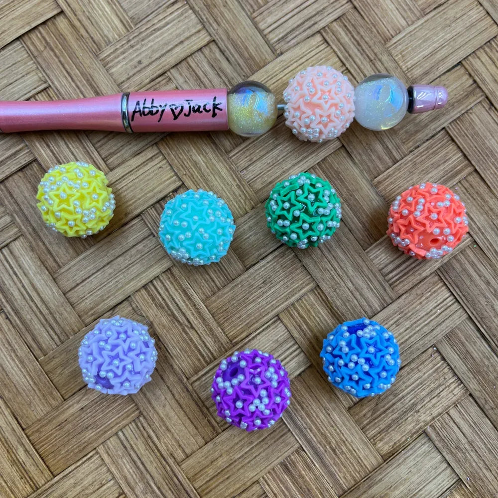 Colorful star-textured pearl sprinkle ball beads in yellow, teal, green, orange, lavender, purple, and blue displayed next to a pink Abby & Jack beadable pen on a woven wood surface
