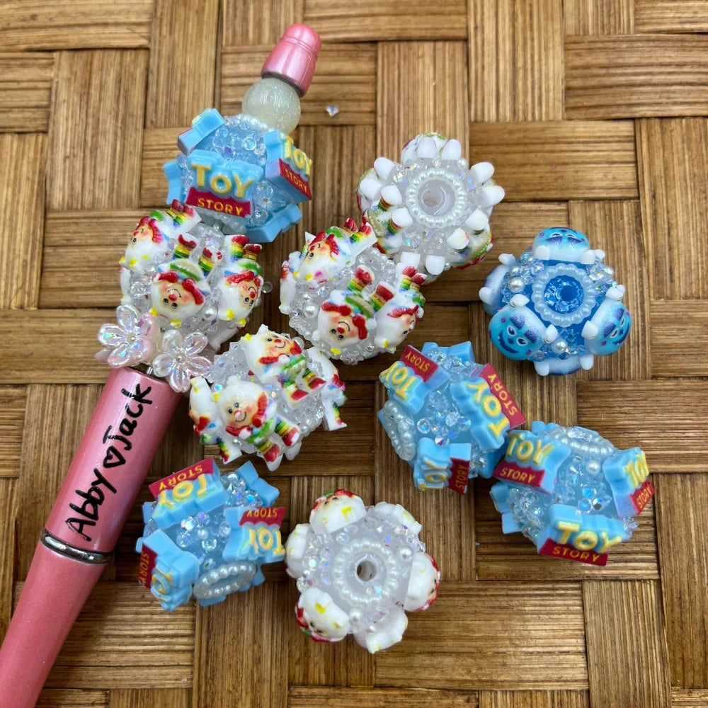 Assorted Toy Story logo and Rainbow Clown character fancy beads with crystal rhinestones and pearl accents in blue and white, displayed on a woven surface next to a decorated pink beadable pen.