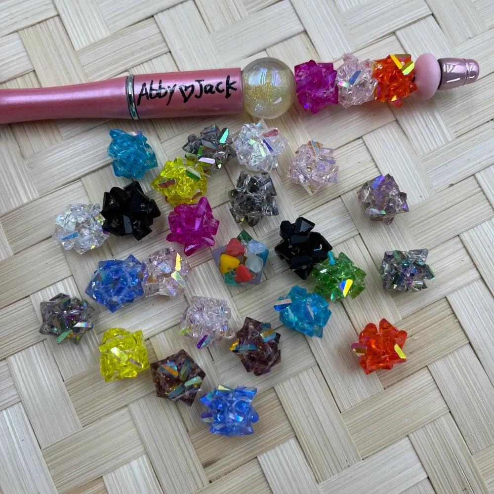 10MM crystal cluster acrylic fancy beads in assorted colors including pink, blue, yellow, black, orange, green and clear with glitter accents displayed next to a beadable pen on a wicker surface.