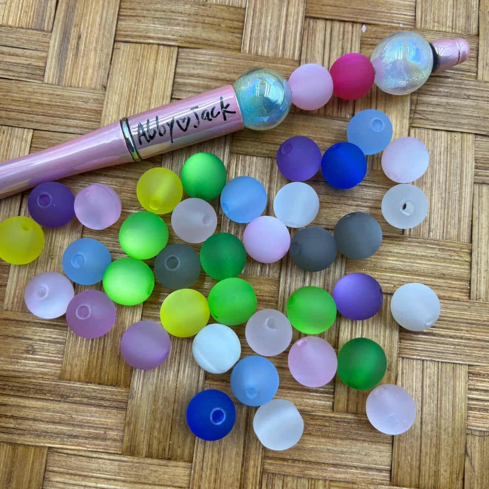 10MM frosted matte multicolor acrylic round beads in assorted colors including green, blue, purple, pink, yellow, and white displayed next to a beadable pen on a wicker surface.