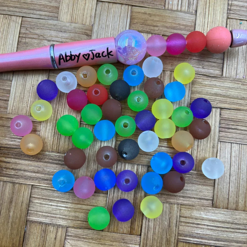10MM frosted matte multicolor acrylic beads in assorted colors including green, blue, purple, yellow, red, and brown, displayed next to a pink beadable pen on a wicker surface.