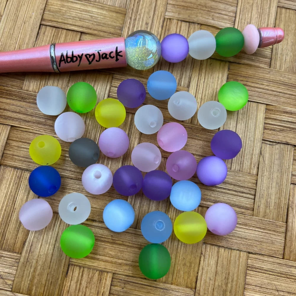 10MM frosted matte rainbow mix acrylic beads in assorted colors including purple, green, yellow, blue, pink, and white, displayed with a beadable pen on a woven surface.