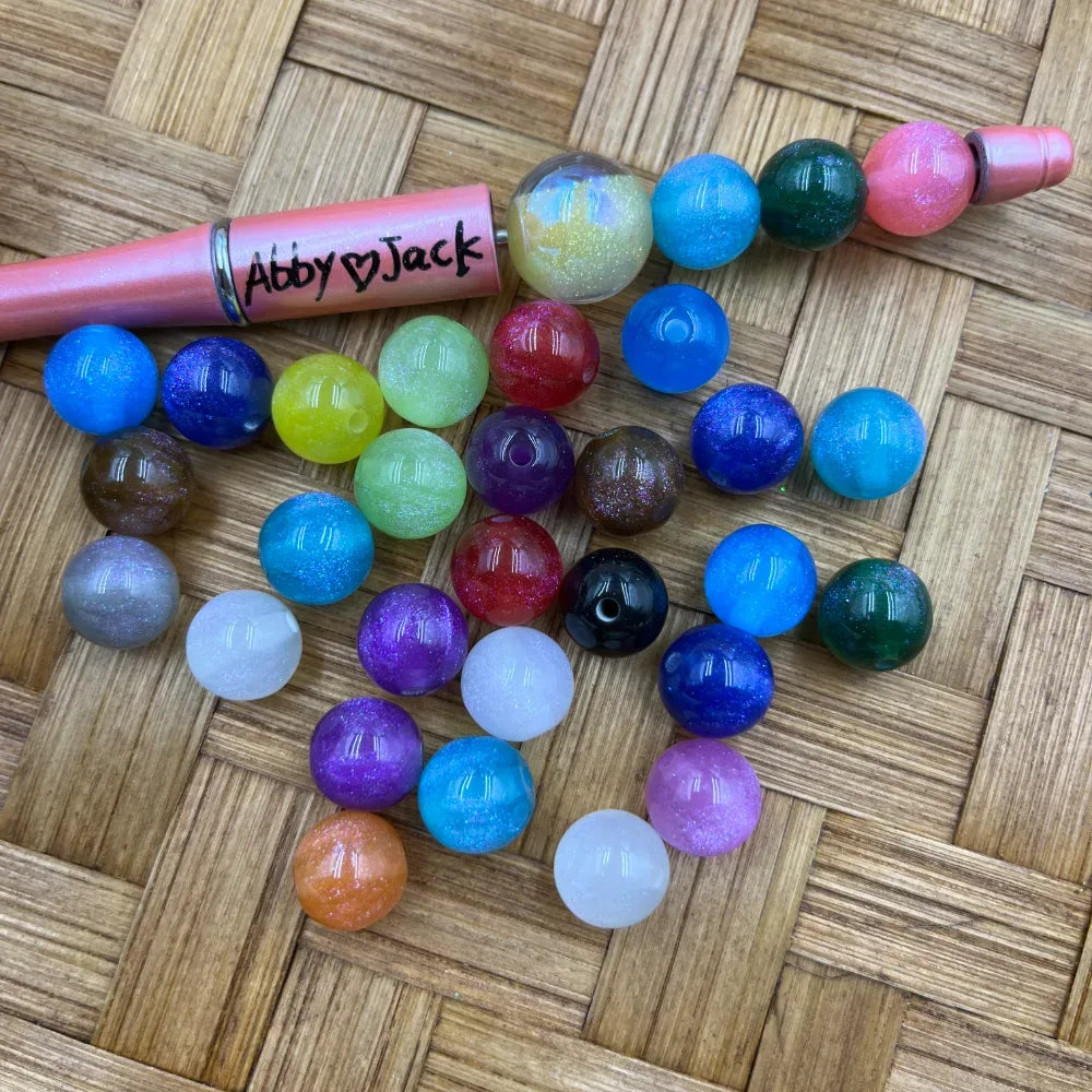10MM multicolor glitter galaxy acrylic beads in assorted colors including blue, purple, green, pink, red, orange, teal, and white, displayed next to a pink beadable pen on a wicker surface.
