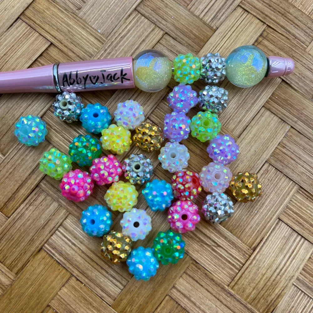 10MM multicolor rhinestone sparkle acrylic beads in pastel pink, blue, yellow, green, purple and silver displayed on a wicker surface next to a pink beadable pen branded Abby Jack.