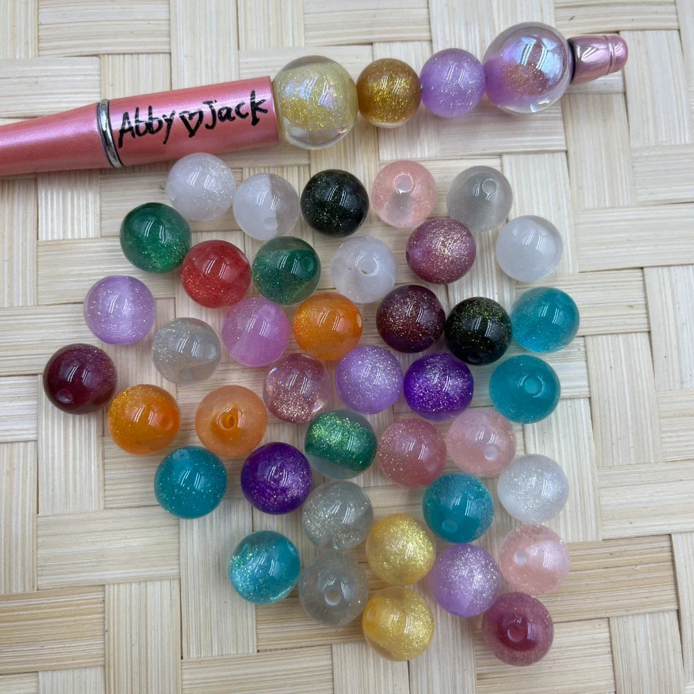 12MM assorted glitter acrylic beads in multiple colors including teal, purple, pink, orange, green, and clear with sparkle finish, displayed next to a beadable pen on a wicker surface.