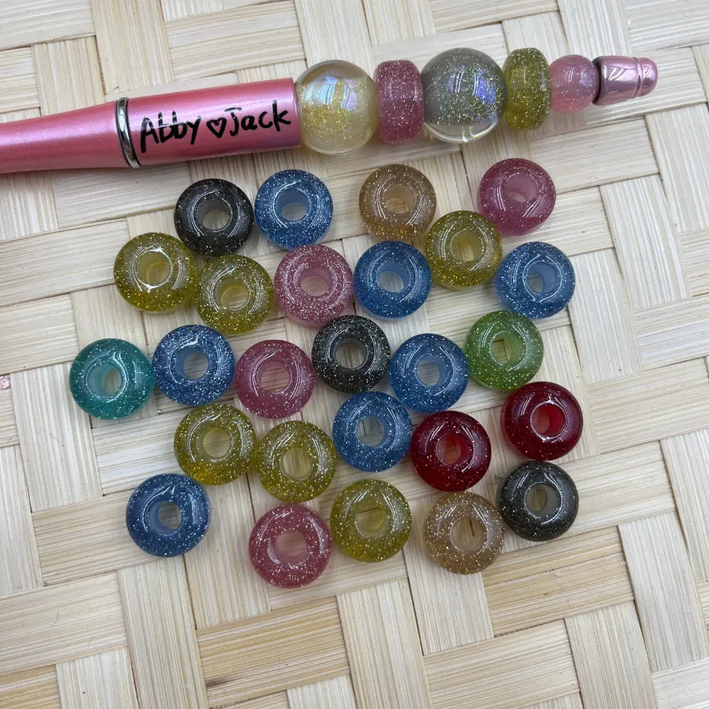 12MM glitter acrylic spacer beads in assorted colors including blue, pink, olive, red, teal, and black with sparkle finish, shown with a beadable pen on a woven surface.