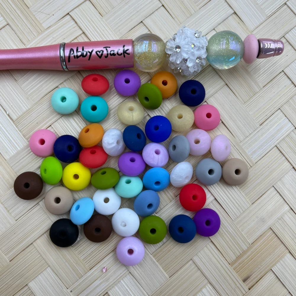 12MM mixed color silicone spacer beads in assorted vibrant colors including pink, blue, green, yellow, red, purple, and more, displayed next to a beaded pen on a wicker surface.