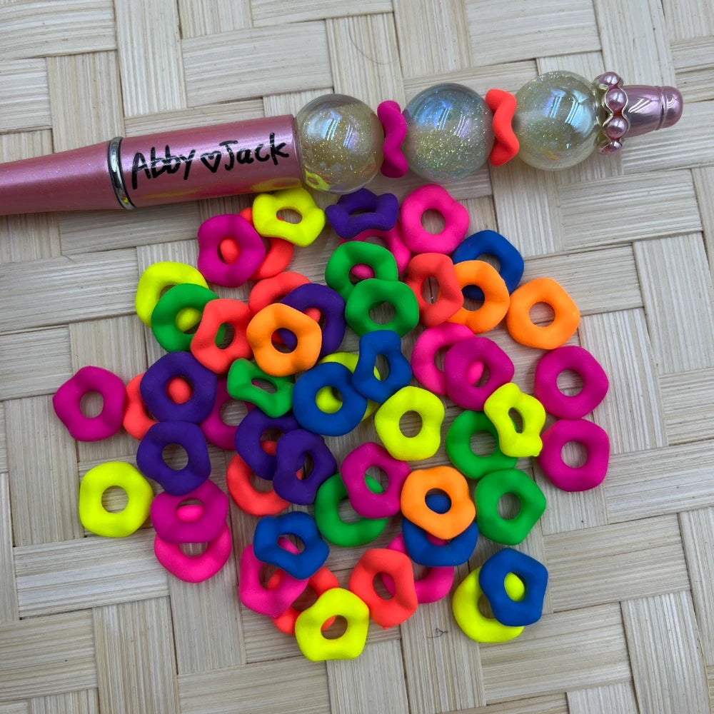 12MM neon rainbow flower ring acrylic spacer beads in hot pink, orange, yellow, green, blue, and purple scattered on a wicker surface next to a beadable pen.