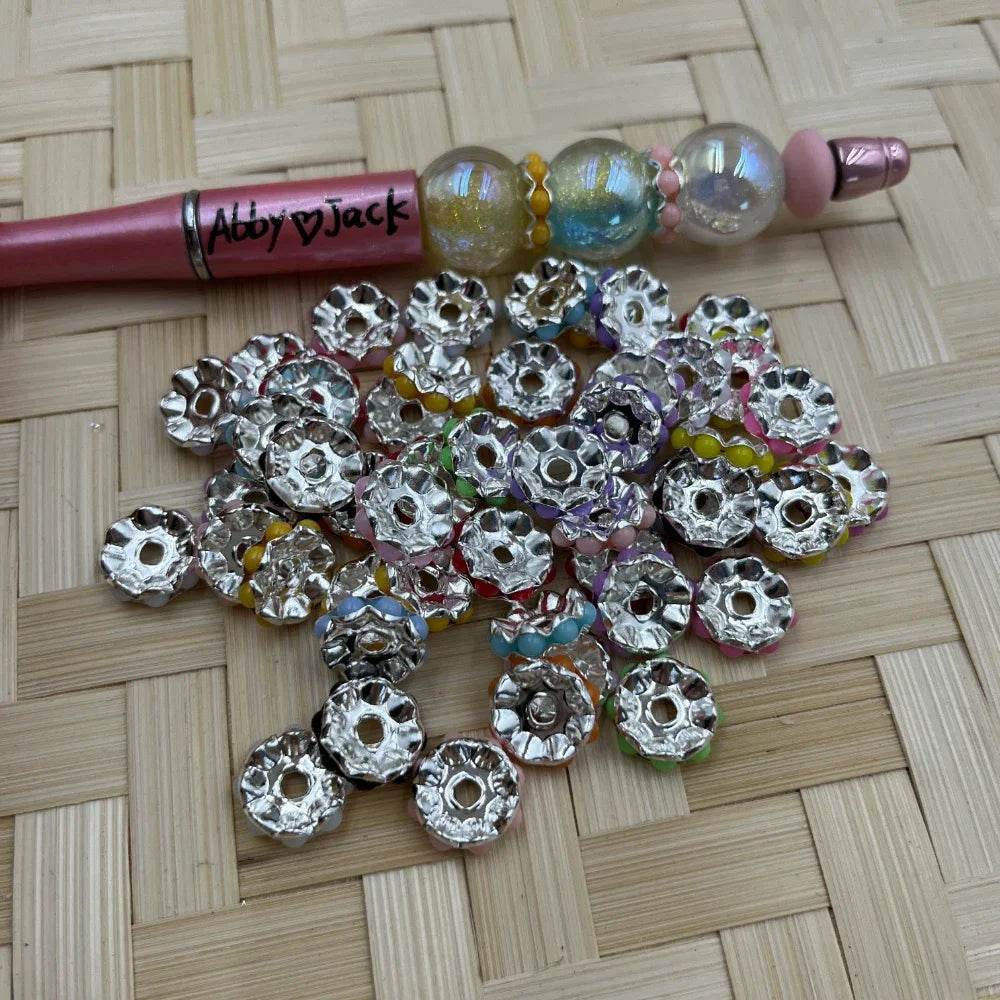 Pile of 12MM silver metal spacer beads with multicolor rhinestone edges beside a pink beadable pen