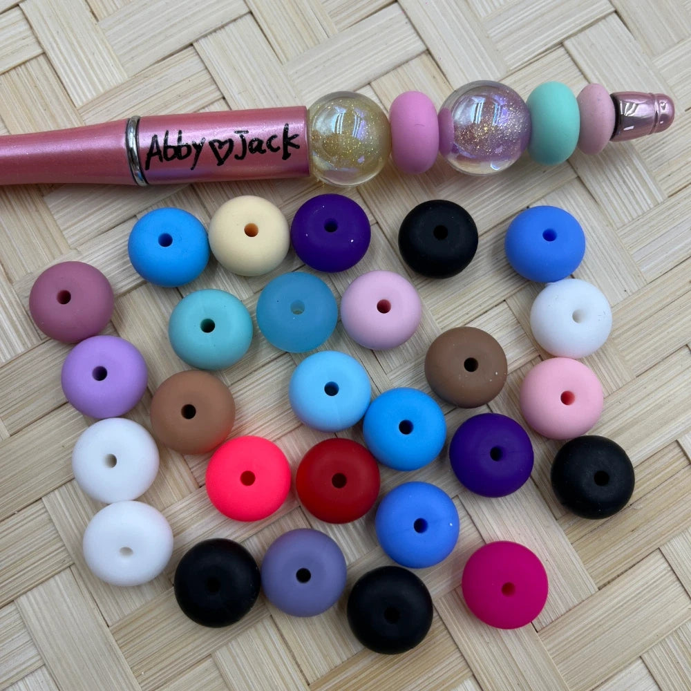 14MM flat round silicone spacer beads in assorted colors including blue, purple, black, pink, red, white, teal, and more, displayed next to a beadable pen on a wicker surface.