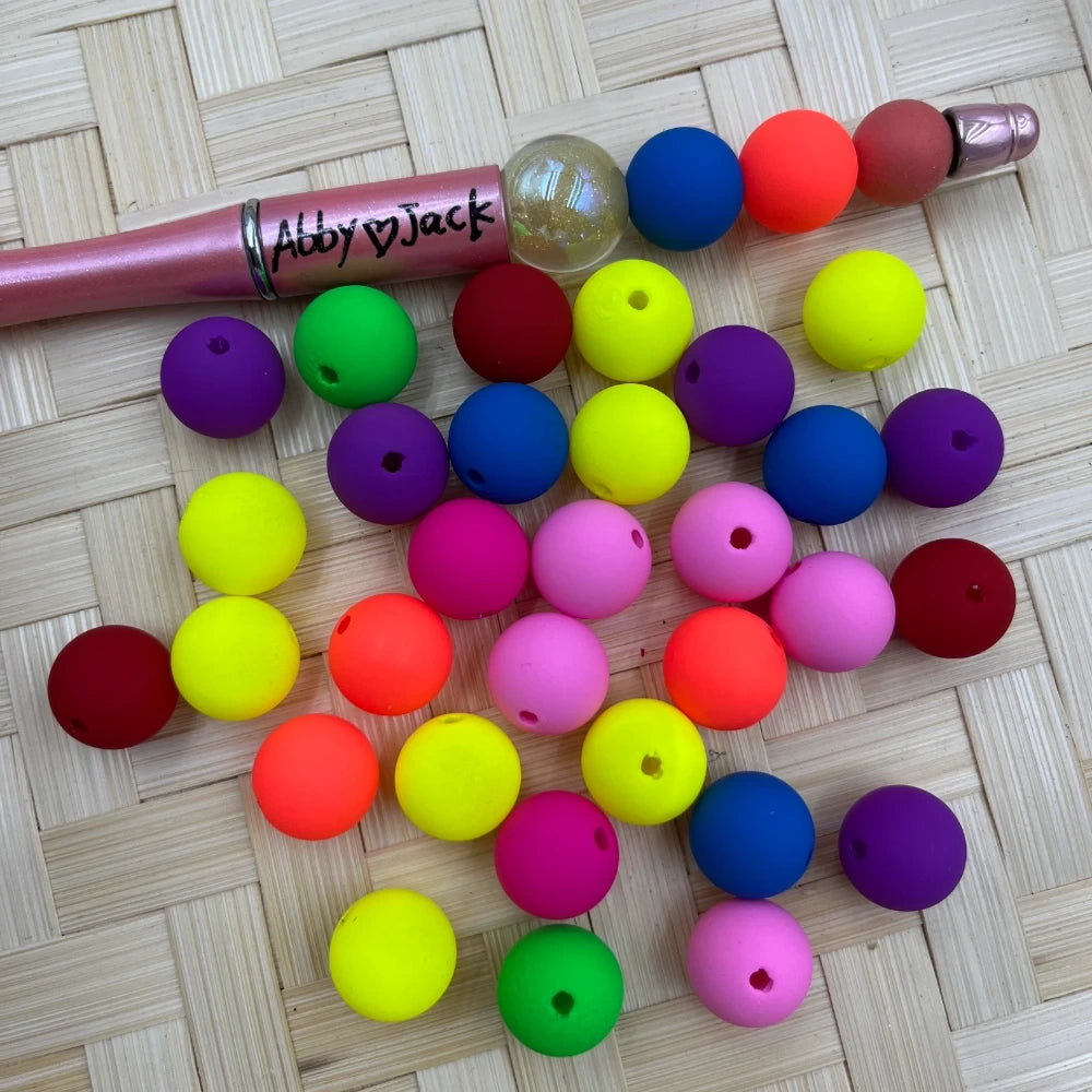 14MM neon rainbow matte acrylic beads in mixed colors including yellow, orange, pink, purple, blue, green and red scattered on a wicker surface next to a beadable pen.