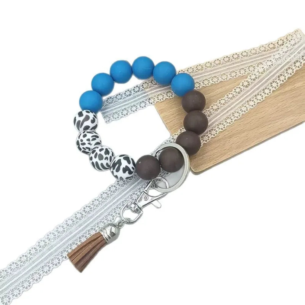 Four silicone bead wristlet keychains featuring cow print and solid colored beads in blue, red, pink, and brown with tassel and lobster clasp hardware on a gray surface. - Blue