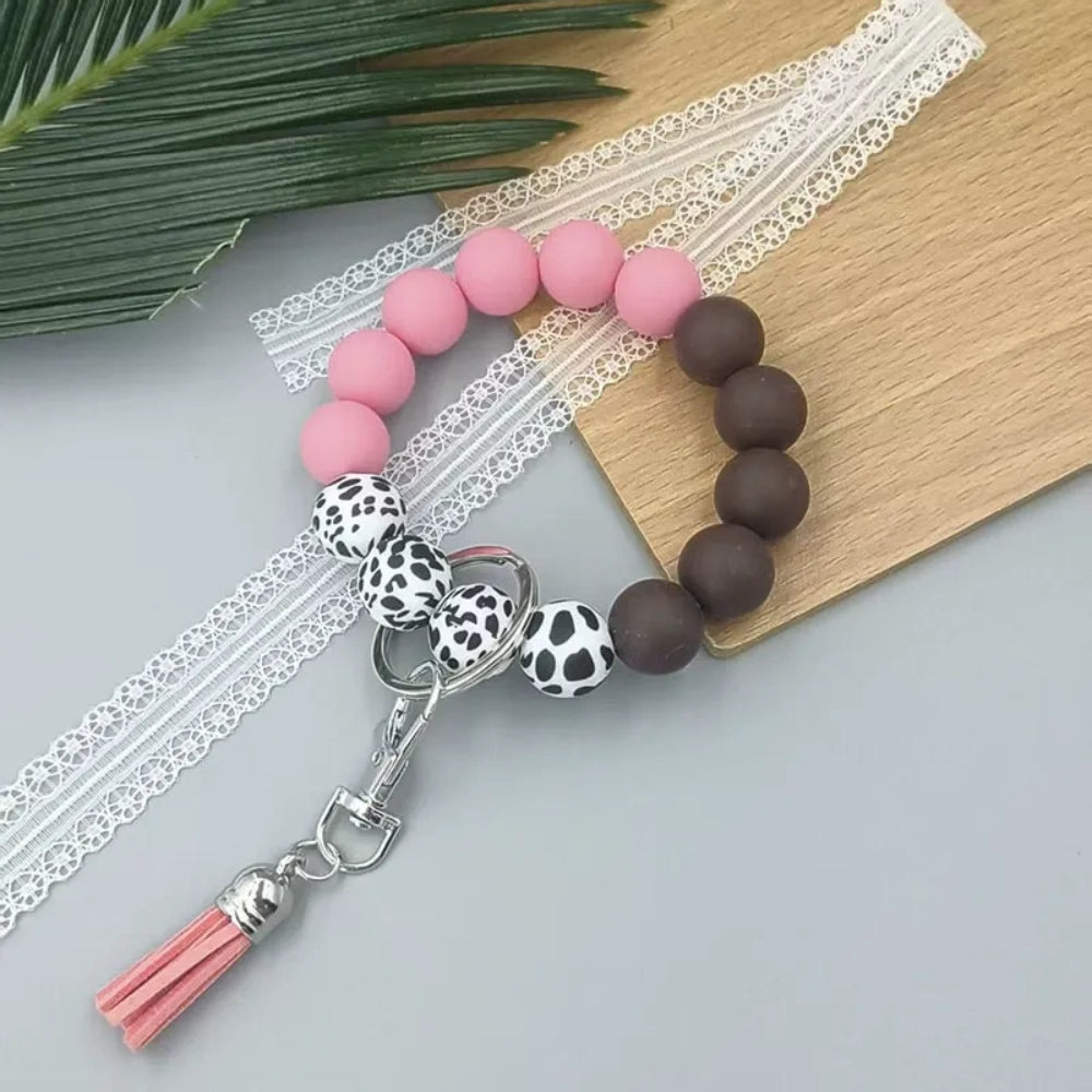 Four silicone bead wristlet keychains featuring cow print and solid colored beads in blue, red, pink, and brown with tassel and lobster clasp hardware on a gray surface. - Pink
