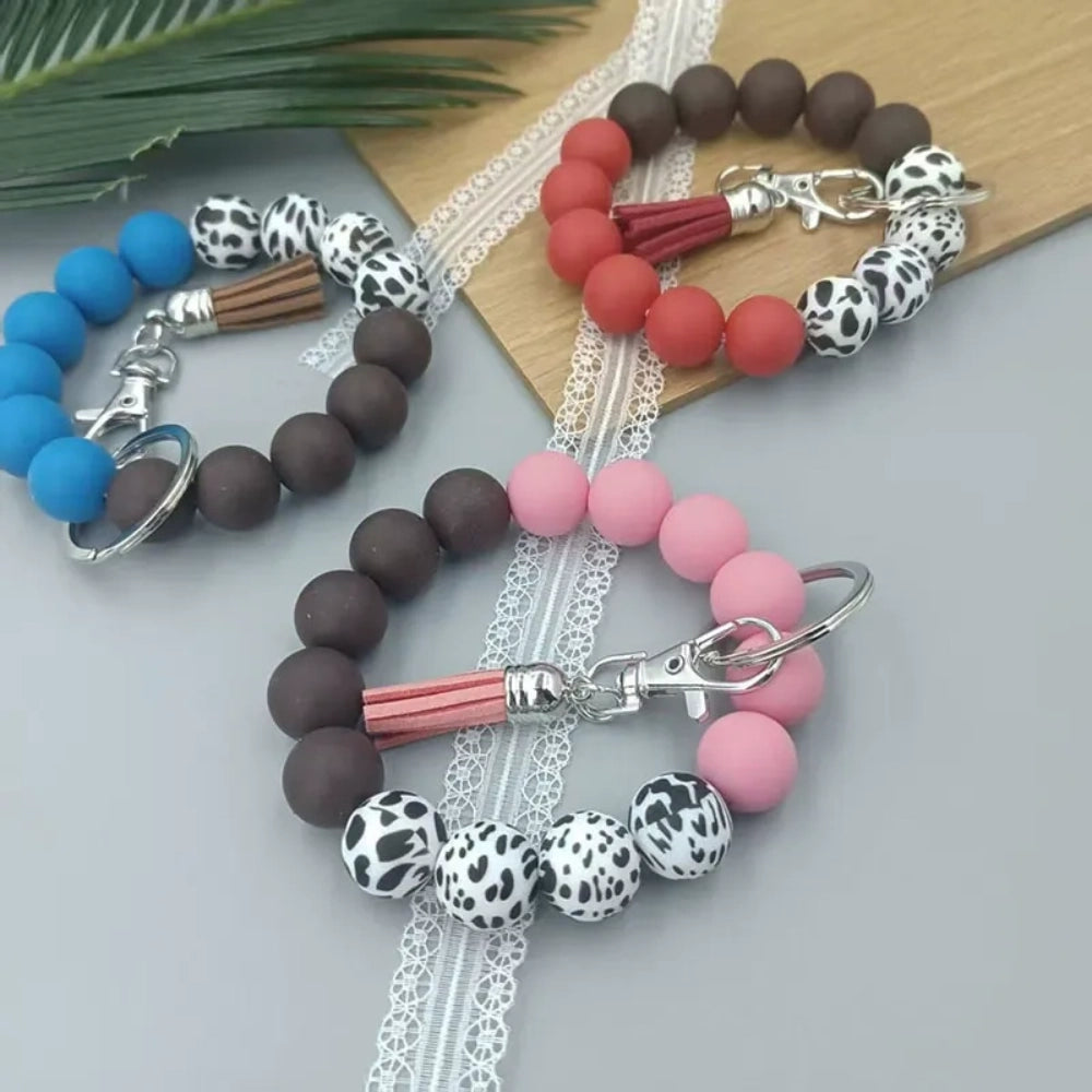 Four silicone bead wristlet keychains featuring cow print and solid colored beads in blue, red, pink, and brown with tassel and lobster clasp hardware on a gray surface. - Random Mixed
