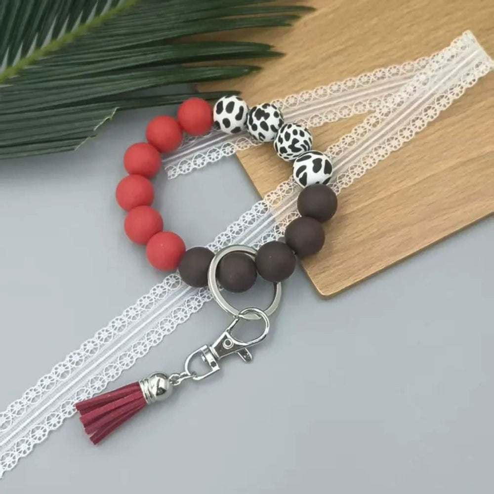 Four silicone bead wristlet keychains featuring cow print and solid colored beads in blue, red, pink, and brown with tassel and lobster clasp hardware on a gray surface. - Red