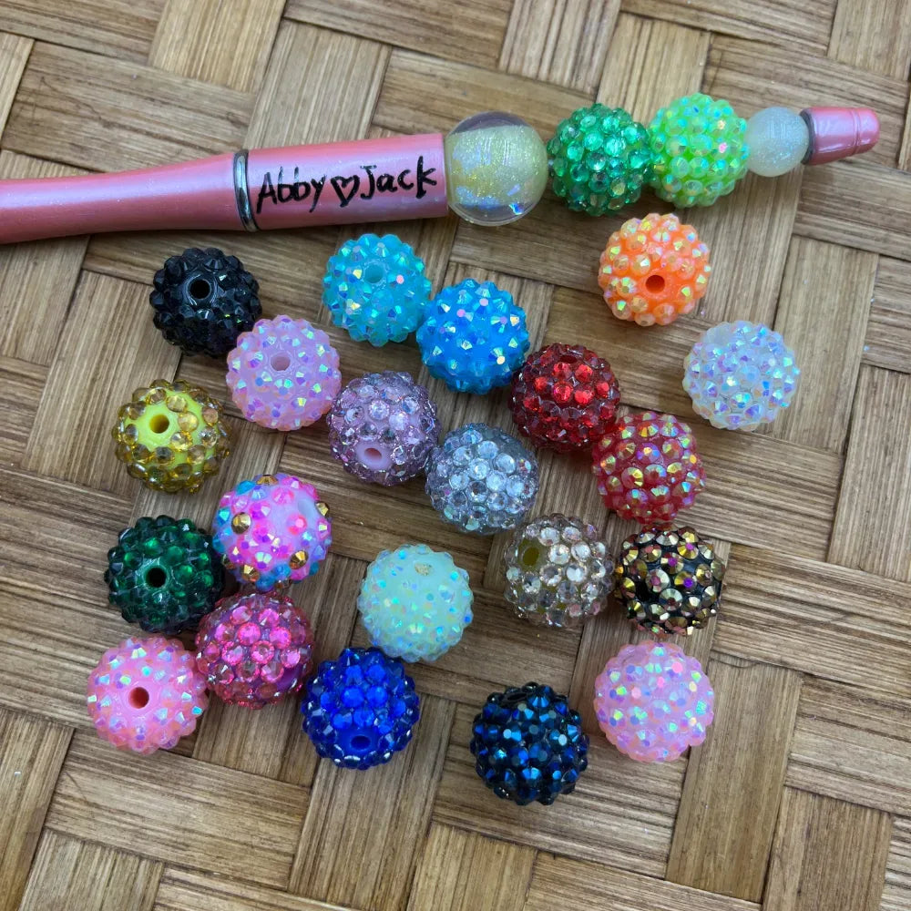 16MM AB rhinestone beads in assorted mixed colors including black, pink, blue, red, green, orange, gold, clear, and white displayed on a wicker surface next to a beadable pen.
