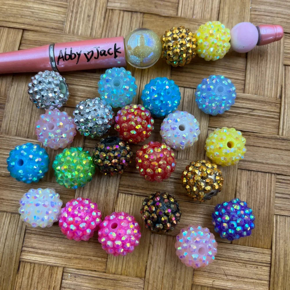 16MM AB rhinestone beads in mixed colors including blue, red, yellow, pink, green, gold, black, purple, and white with iridescent sparkle finish, shown next to a beadable pen on a wicker surface.