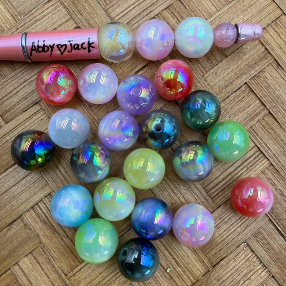 16MM iridescent aurora AB acrylic beads in mixed colors including white, green, black, red, pink, and purple with rainbow holographic shimmer finish, displayed on a woven surface next to a beadable pen.