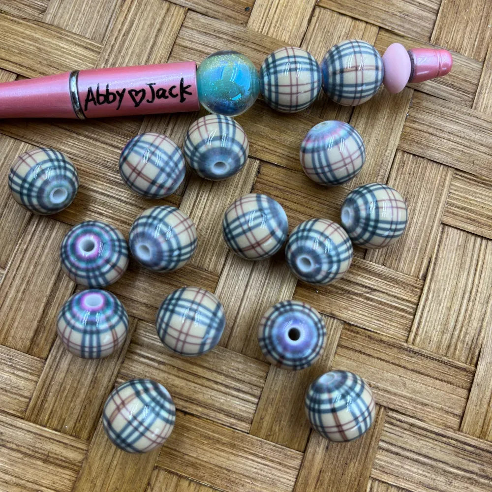 16MM tan and black plaid print acrylic beads scattered on wicker surface next to a custom beadable pen labeled Abby Jack, showing iridescent hole detail on round beads.
