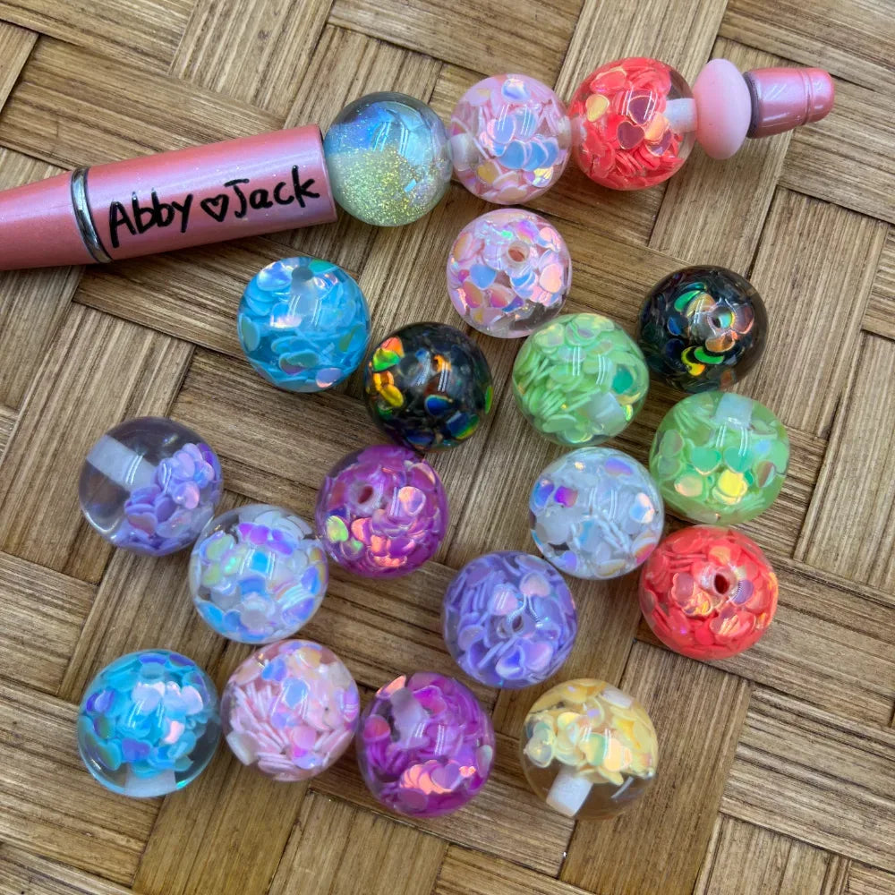 16MM iridescent confetti heart glitter acrylic beads in assorted colors including blue, pink, purple, green, black, red, and yellow displayed on a wicker surface next to a beadable pen.