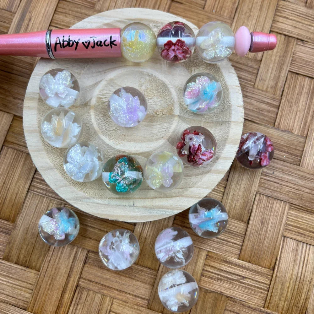 16MM clear acrylic round beads encasing dried flowers and butterfly embellishments in white, red, pink, teal, and iridescent glitter finishes, displayed on a wooden tray with a beadable pen.