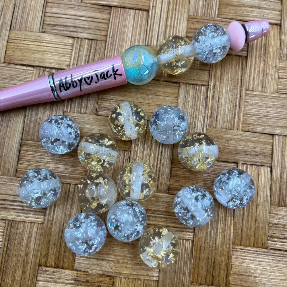16MM clear acrylic round beads with gold and silver foil flakes inside crackle finish, displayed alongside a pink beadable pen on a woven bamboo surface.