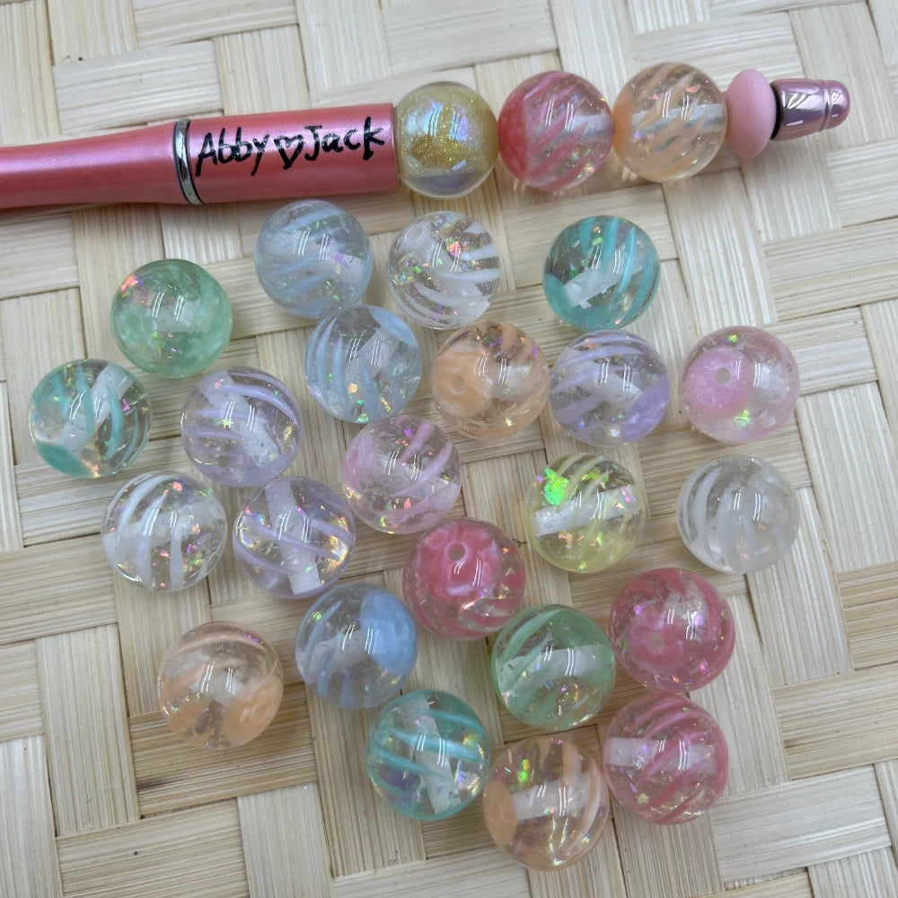 16MM pastel iridescent glitter swirl acrylic beads in pink, green, peach, and clear shown with a beadable pen on a wicker surface