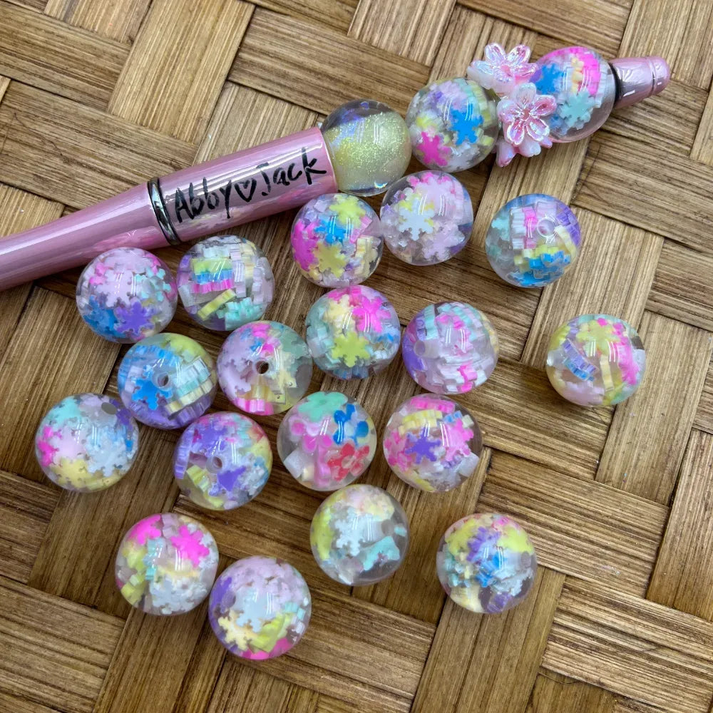 16MM clear acrylic round beads filled with pastel rainbow confetti snowflake and star shapes, displayed alongside a pink beadable pen on a wicker surface.