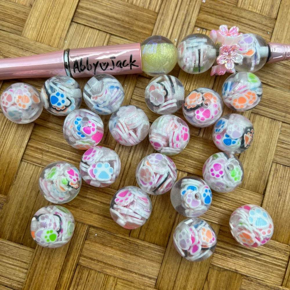 16MM clear round acrylic beads filled with colorful paw print confetti pieces in pink, blue, orange, green, and purple, displayed on a wicker surface next to a pink beadable pen labeled Abby Jack.
