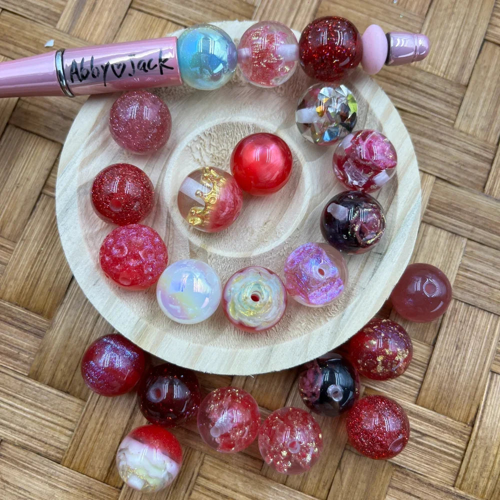 Assorted 16MM red glitter galaxy mix acrylic beads in red, cranberry, iridescent, and clear gold foil finishes displayed on a wooden tray with a pink beadable pen.