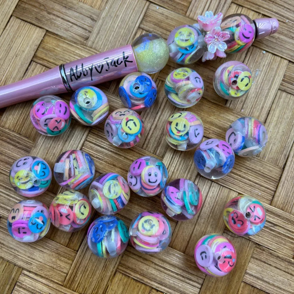 16MM clear acrylic round beads filled with colorful smiley face emoji confetti slices in pastel pink, yellow, blue, and purple, displayed on wicker surface next to a pink beadable pen.