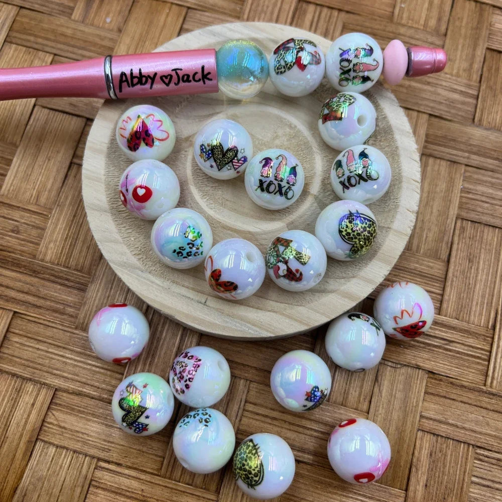 16MM iridescent white acrylic beads featuring Valentine XOXO gnomes, leopard print hearts, and butterfly designs arranged on a wooden tray with a pink beadable pen labeled Abby Jack.