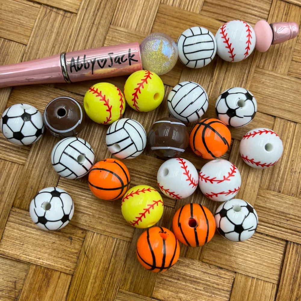 16MM sports ball acrylic beads in soccer, baseball, basketball, softball, and volleyball designs scattered on a woven surface next to a beadable pen.
