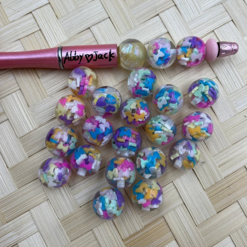 16MM clear acrylic round beads filled with colorful pastel butterfly and star confetti shapes, shown with a pink beadable pen on a woven background.
