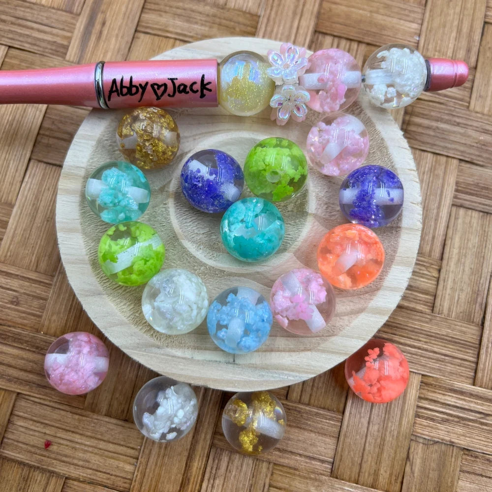 16MM crushed crystal galaxy acrylic beads in assorted colors including teal, purple, pink, orange, green, white, and gold displayed on a wooden tray with a pink beadable pen labeled Abby and Jack