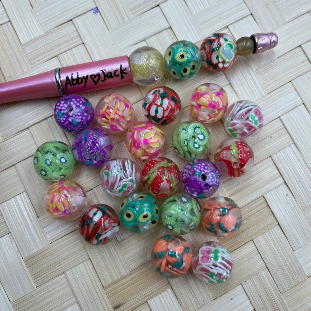 16MM fruit and floral confetti acrylic beads featuring kiwi, mushroom, flower and mixed pattern designs in colorful transparent resin, displayed with a pink beadable pen on a wicker surface.