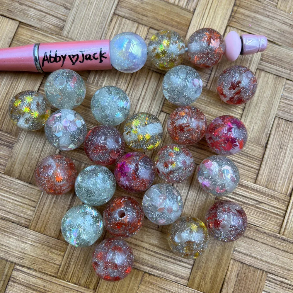 16MM round acrylic beads with galaxy glitter, holographic star confetti, and red pink iridescent flakes displayed on woven surface next to a beadable pen labeled Abby Jack.