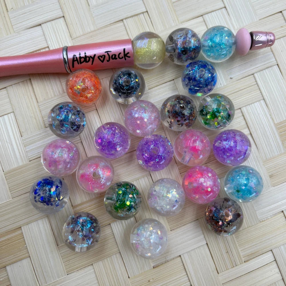 16MM clear acrylic round beads filled with colorful glitter and confetti in assorted colors including pink, purple, blue, orange, and green, displayed on a wicker surface next to a pink beadable pen.